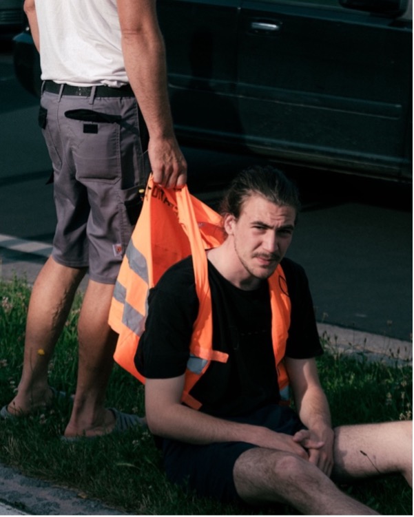 Das Bild zeigt Valentin Bast erschöpft auf dem Boden sitzend am Straßenrand nach einer Klebeaktion. Ein Mann packt ihn n der Warnweste.
